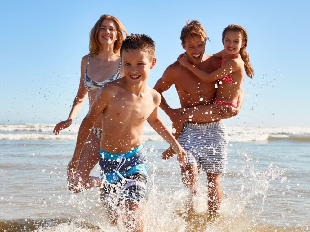 Family of four, happy and running in the ocean waves on a sunny beach.
